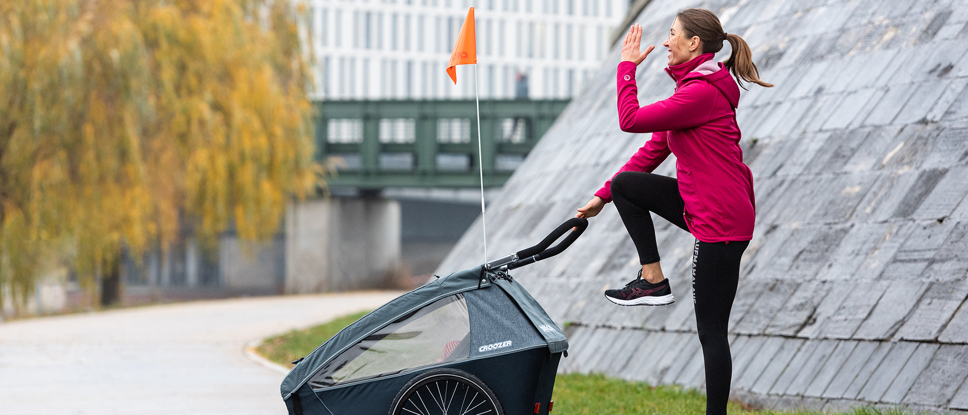 Woman in pink jacket stretching with one leg raised holding the Croozer kid trailer in urban setting.